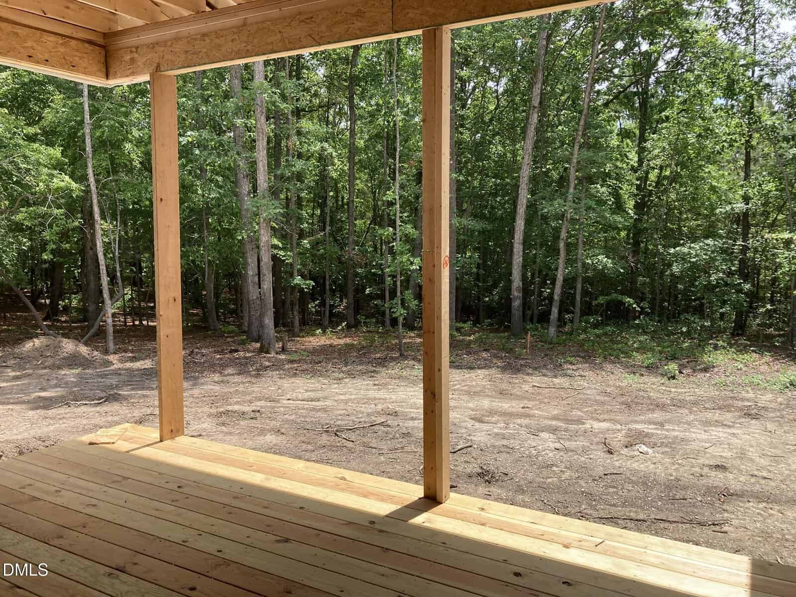 A wooden porch with a roof overlooks a dirt yard and a dense forest of tall green trees on a sunny day.