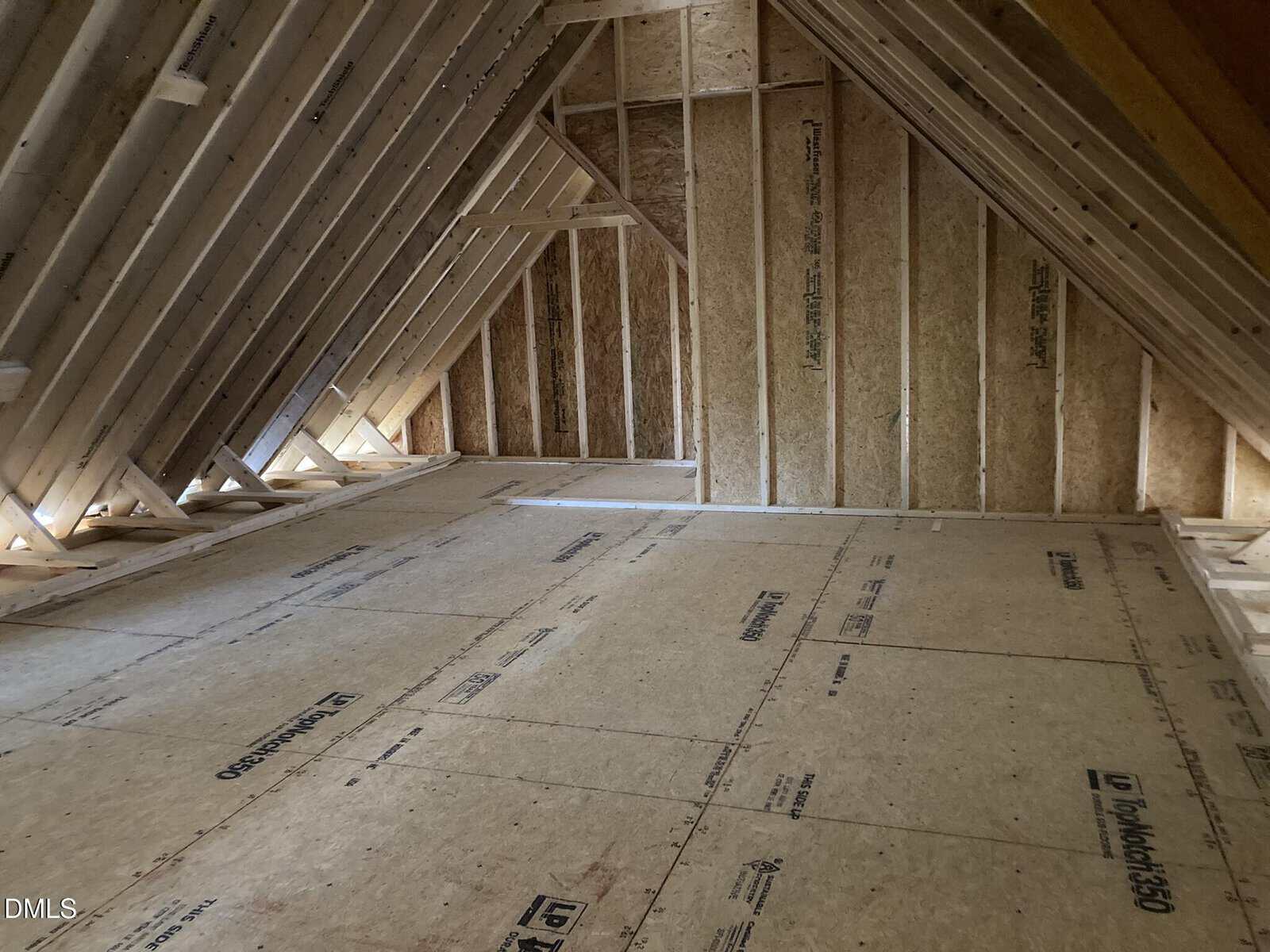 Unfinished attic space with exposed wooden framing and plywood floor. Sunlight enters through gaps in the roof structure. The area is empty and appears under construction.