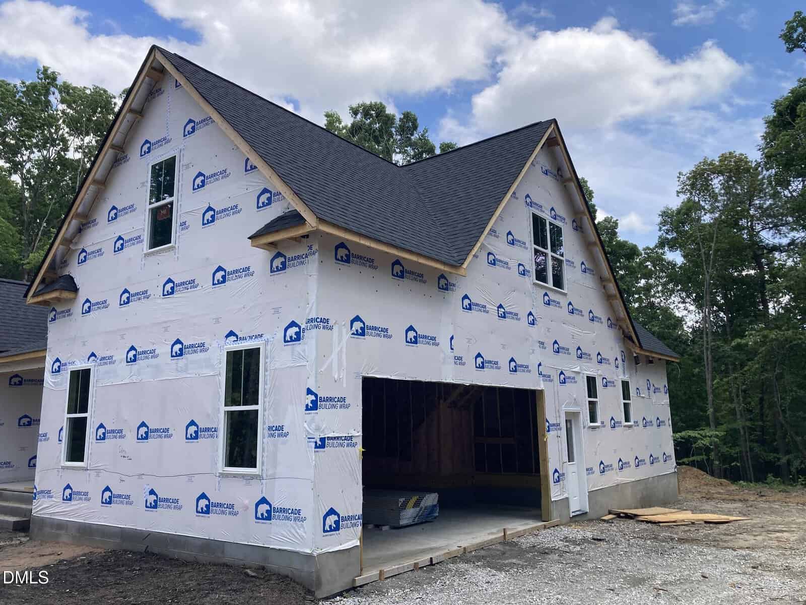 A house under construction with white weather-resistant wrap, black roof shingles, exposed wood framing in the garage, and surrounded by trees and gravel.