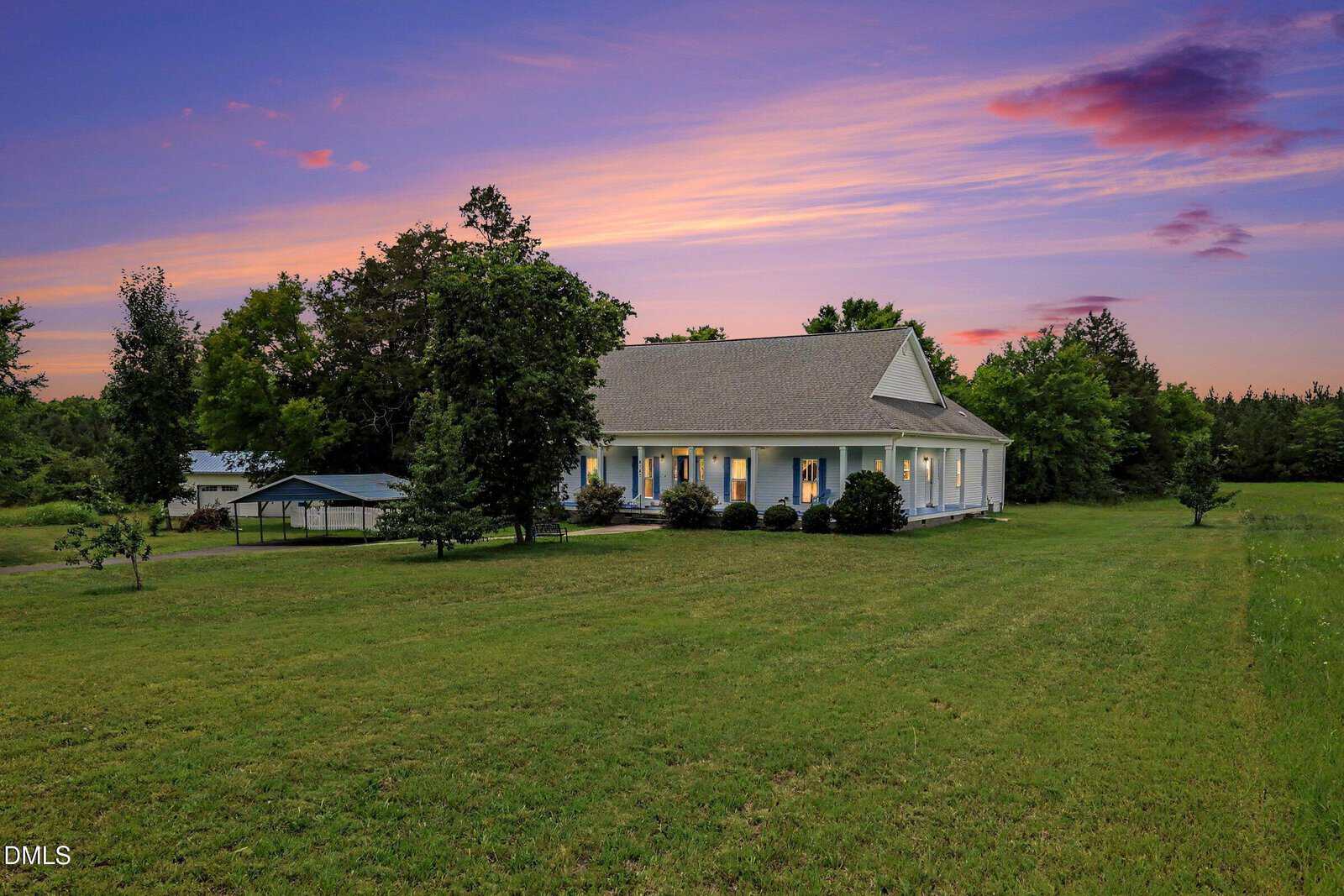 A white house with a porch sits on a large grassy lawn with trees, under a colorful sunset sky with pink and purple clouds.