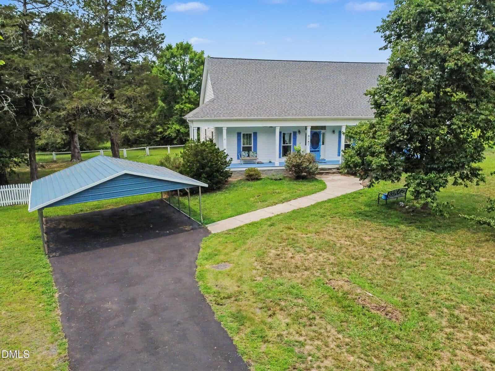 Single-story house with a covered front porch, blue accents, a curved walkway, and a detached carport on a spacious green lawn with trees and a white fence in the background.