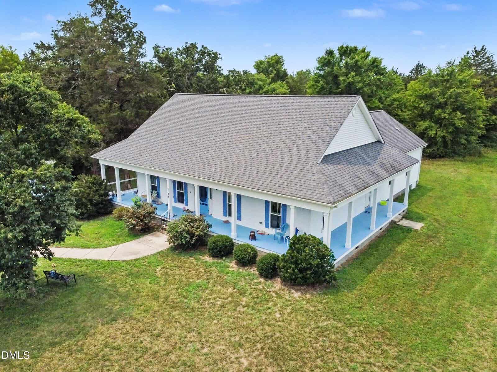 A large white house with a gray roof and blue accents sits gracefully at 9187 Cornwall Road in Oxford, surrounded by green grass, bushes, and trees. The home features a covered front porch and a curved sidewalk leading to the entrance.