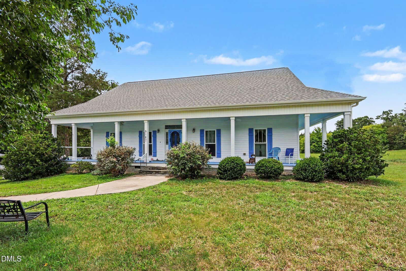 A light blue house with a large wraparound porch, white columns, and a sloped roof, surrounded by bushes and a grassy yard under a sunny, blue sky.