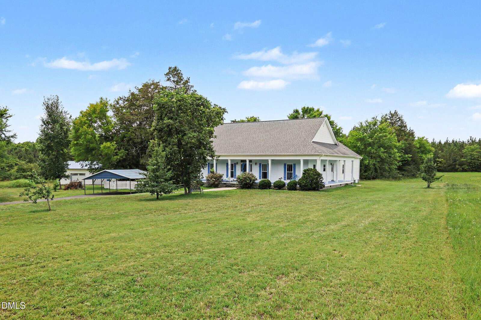 A white house with a large front porch sits on a spacious grassy lawn, surrounded by trees and open sky. A small carport and shed are visible to the left of the house.