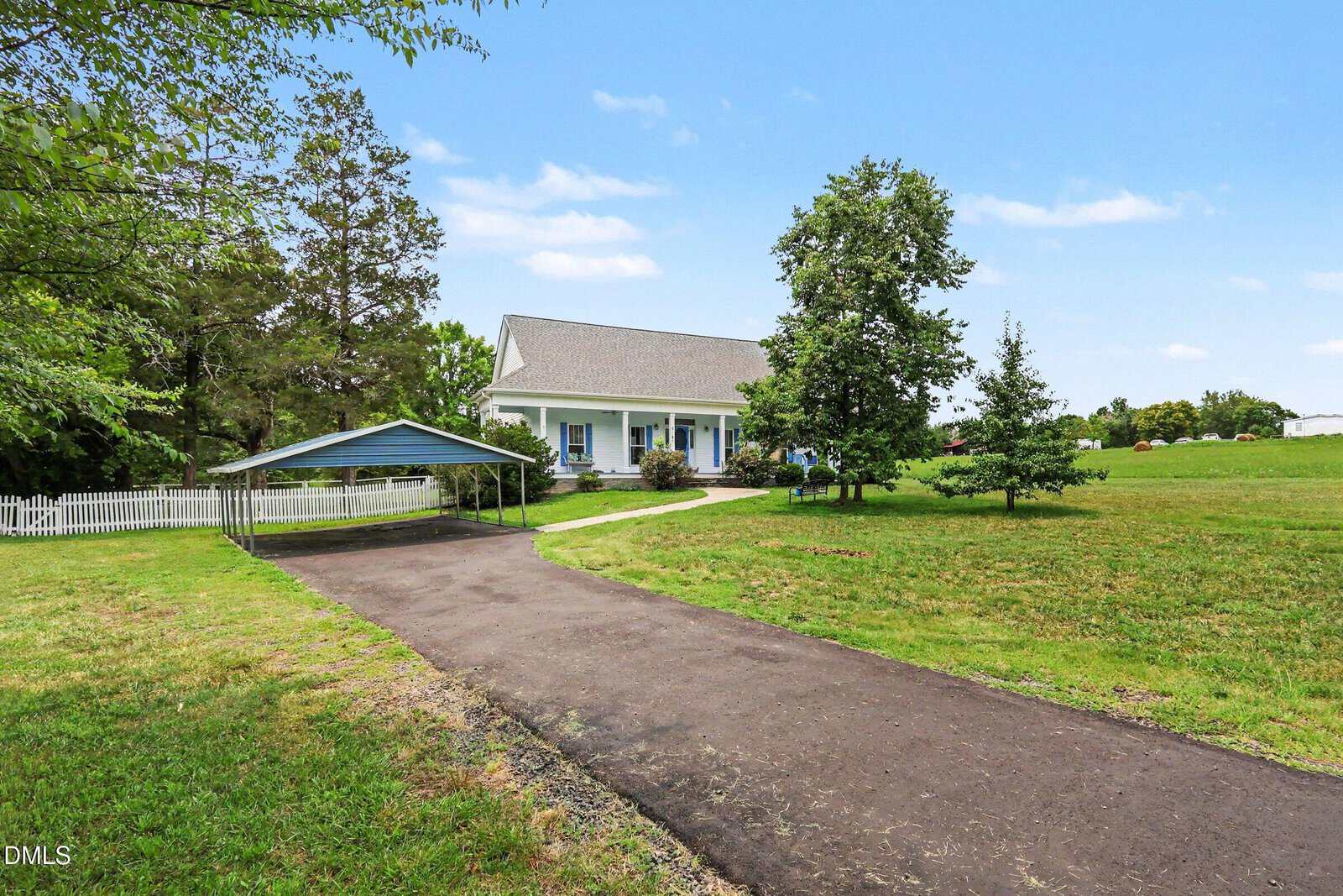 A paved driveway leads to a white house with a porch and a carport, surrounded by green lawn, trees, and a white picket fence under a blue sky.