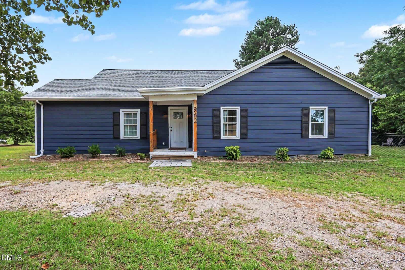 Single-story house with blue siding, white trim, and a gray shingle roof. There are three windows with black shutters, a small front porch with wooden posts, and some bushes in the front yard. Grass and a gravel area are visible.