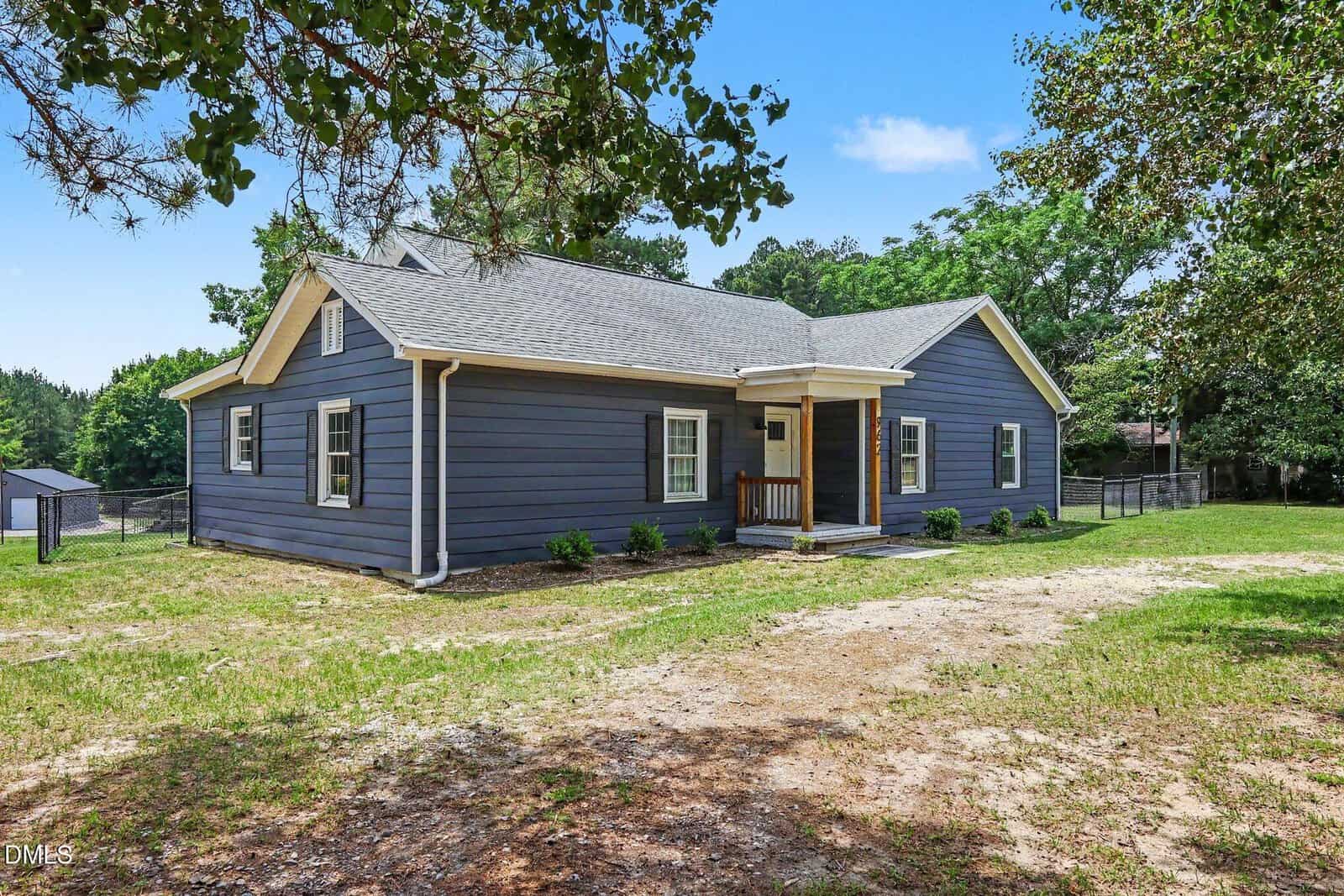 Single-story house with dark blue siding, white trim, a small covered entry porch, fenced backyard, and surrounded by grass and trees under a clear blue sky.
