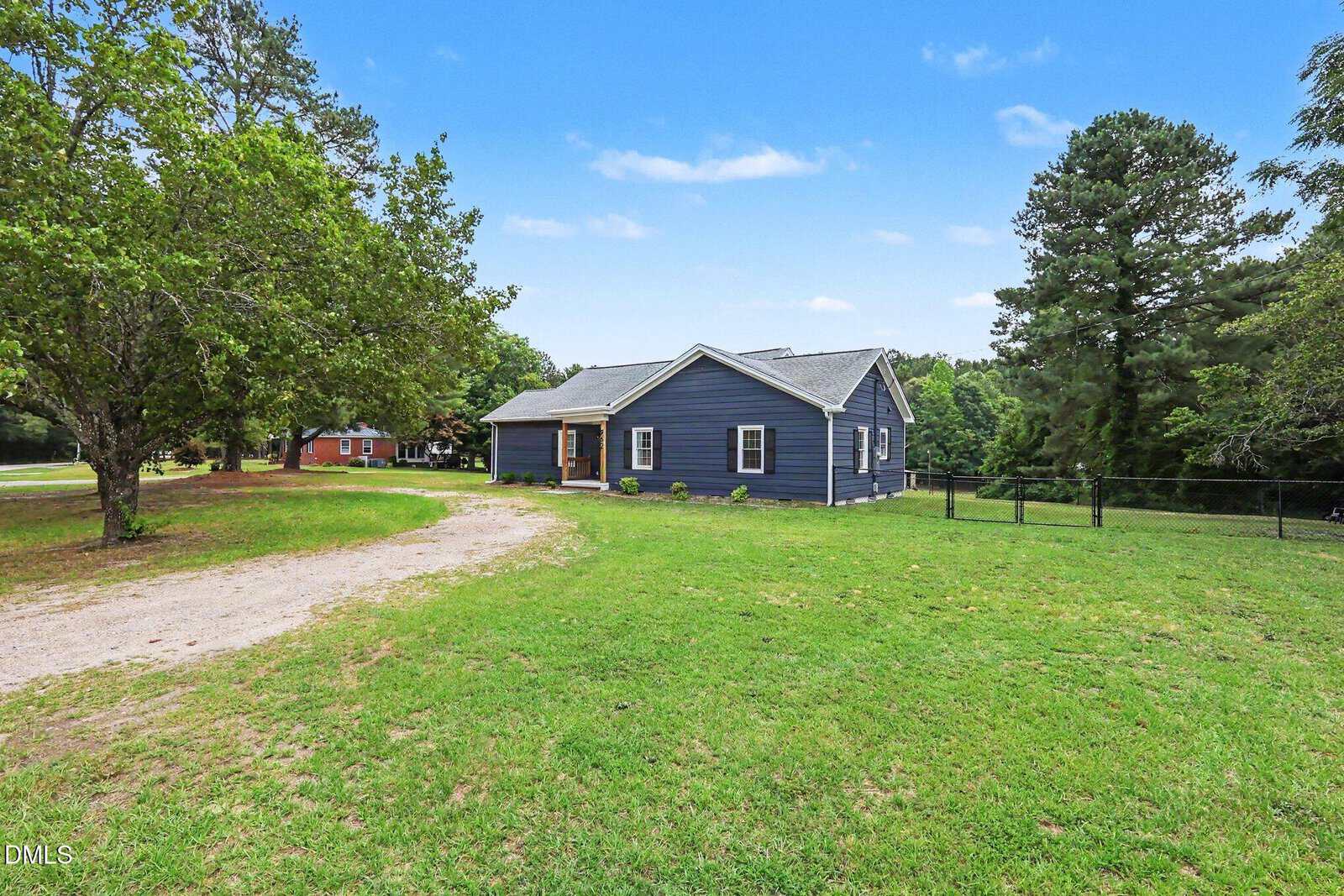 A blue single-story house with a gray roof sits on a spacious grassy yard at 962 Laurel Mill-Centerville Road, Louisburg, surrounded by trees and a dirt driveway, under a clear blue sky.