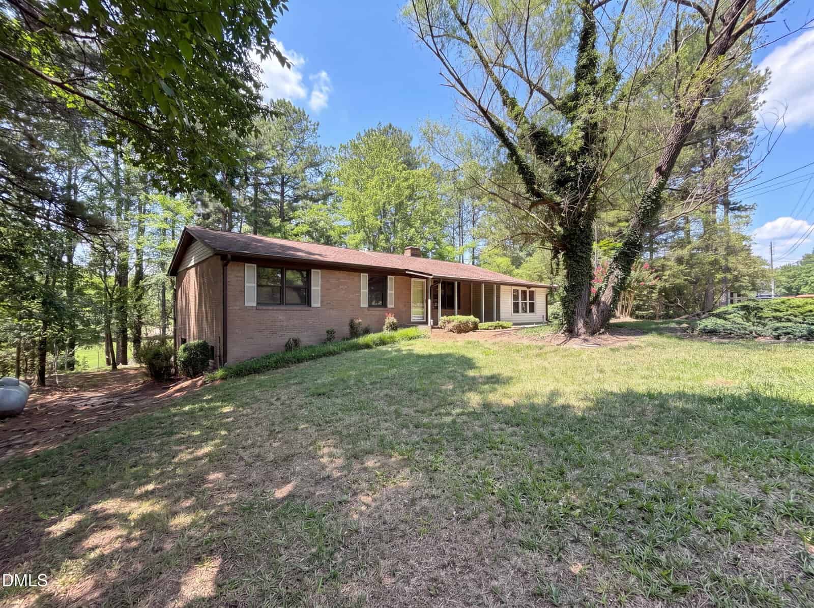 Single-story brick house with a sloped roof, large windows, and a grassy front yard surrounded by tall trees under a blue sky with scattered clouds, ideally situated at 2560 Mangum Avenue in tranquil Creedmoor.