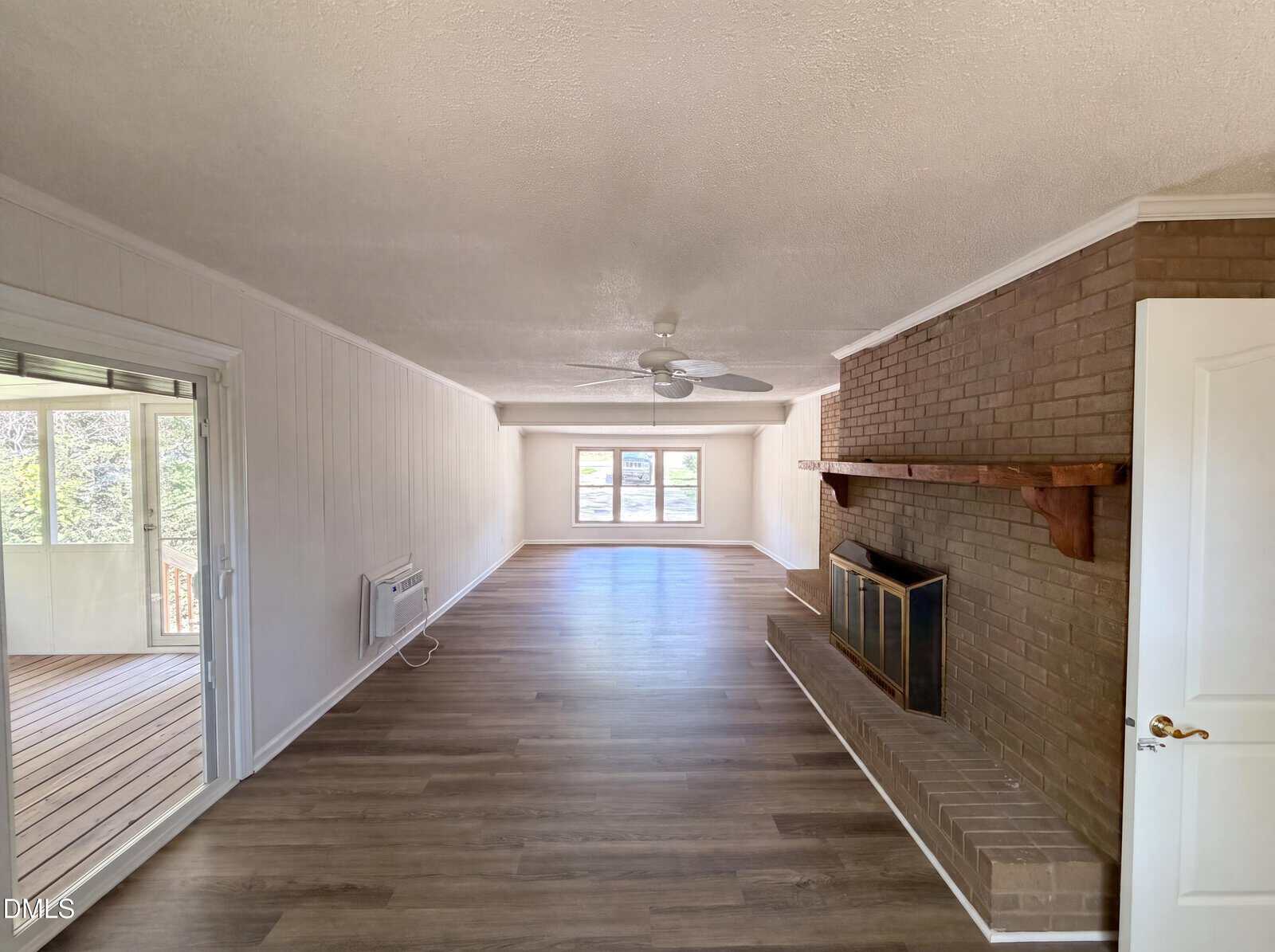 Long living room with wood flooring, a brick fireplace and mantel on the right, white walls, a ceiling fan, and large windows at the far end letting in natural light. A sliding glass door leads to a sunroom on the left.