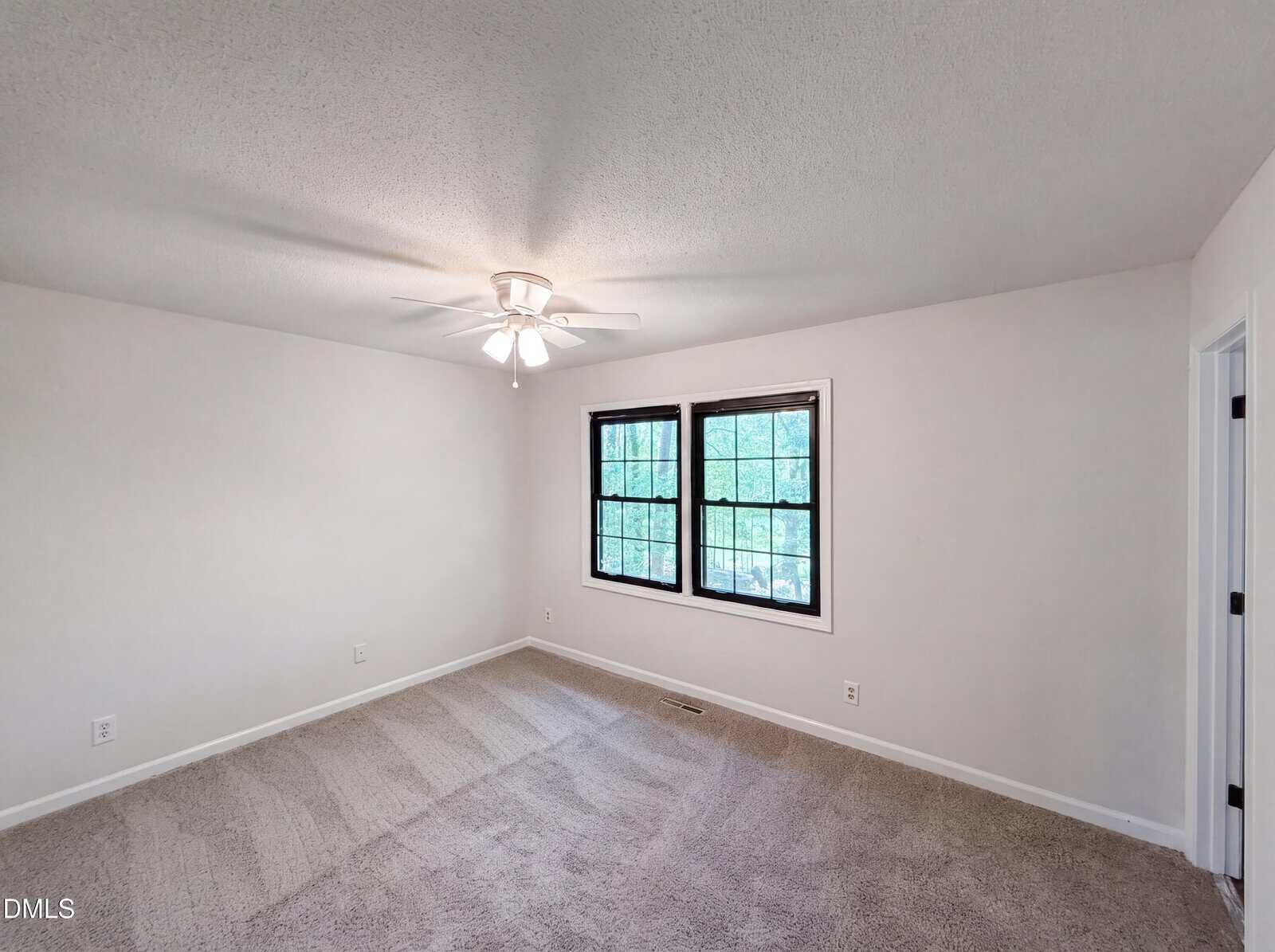 Empty room with beige carpet, white walls, a ceiling fan with light, and two large windows letting in natural light. Doorway is visible on the right.