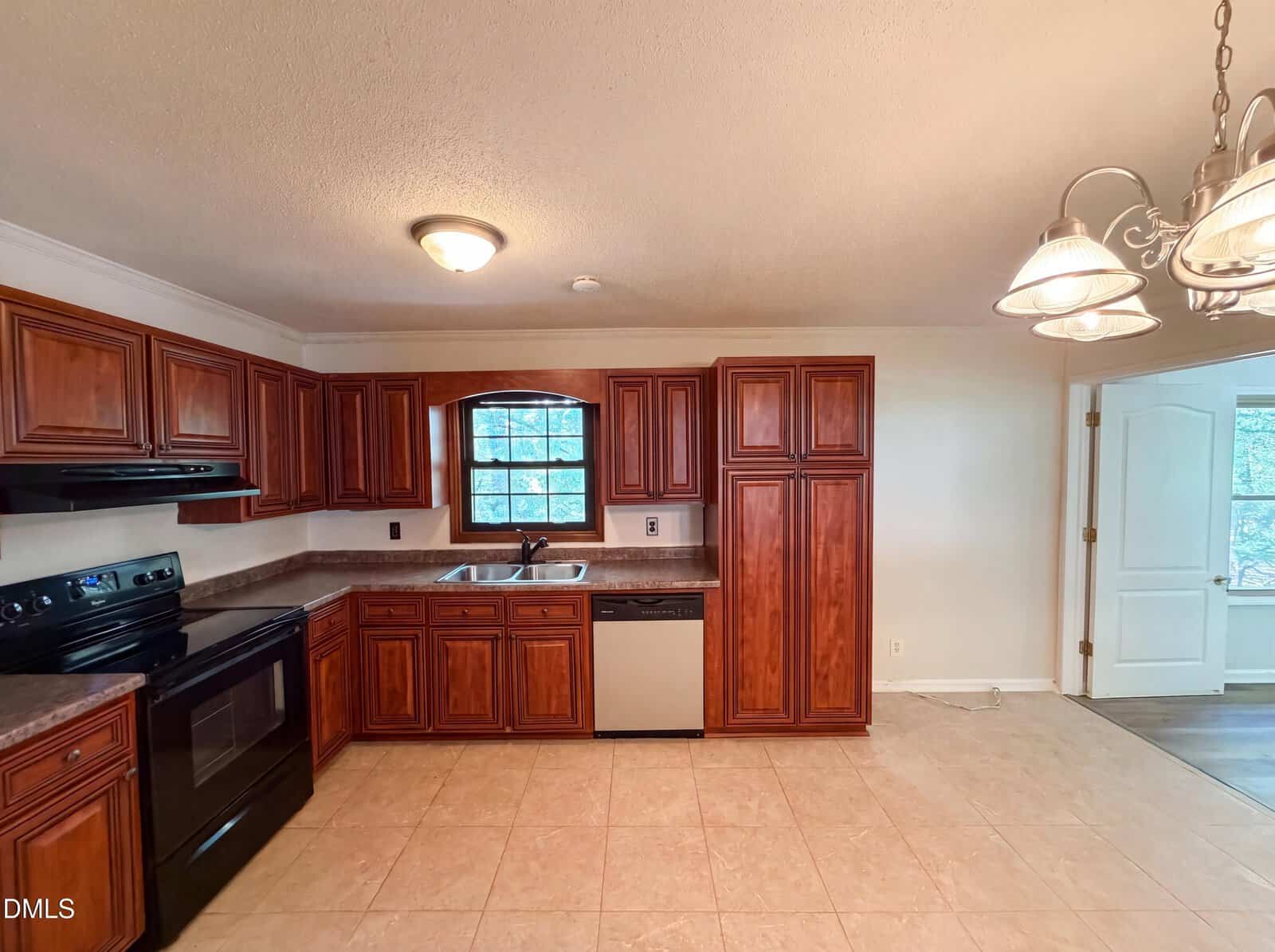 Kitchen with brown wooden cabinets, black appliances, stainless steel dishwasher, tile floor, overhead lighting, and a window above the sink. A chandelier hangs above the dining area on the right. Double doors are partially visible.