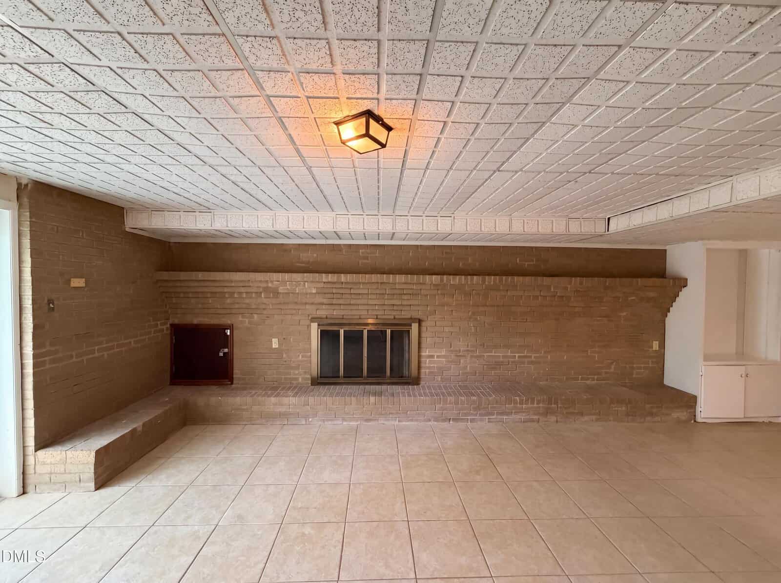A spacious room with tan tile flooring, brick walls, a built-in fireplace, a small red cabinet, and a white light fixture on a tiled ceiling. There is a step-up ledge and built-in shelves along one wall.