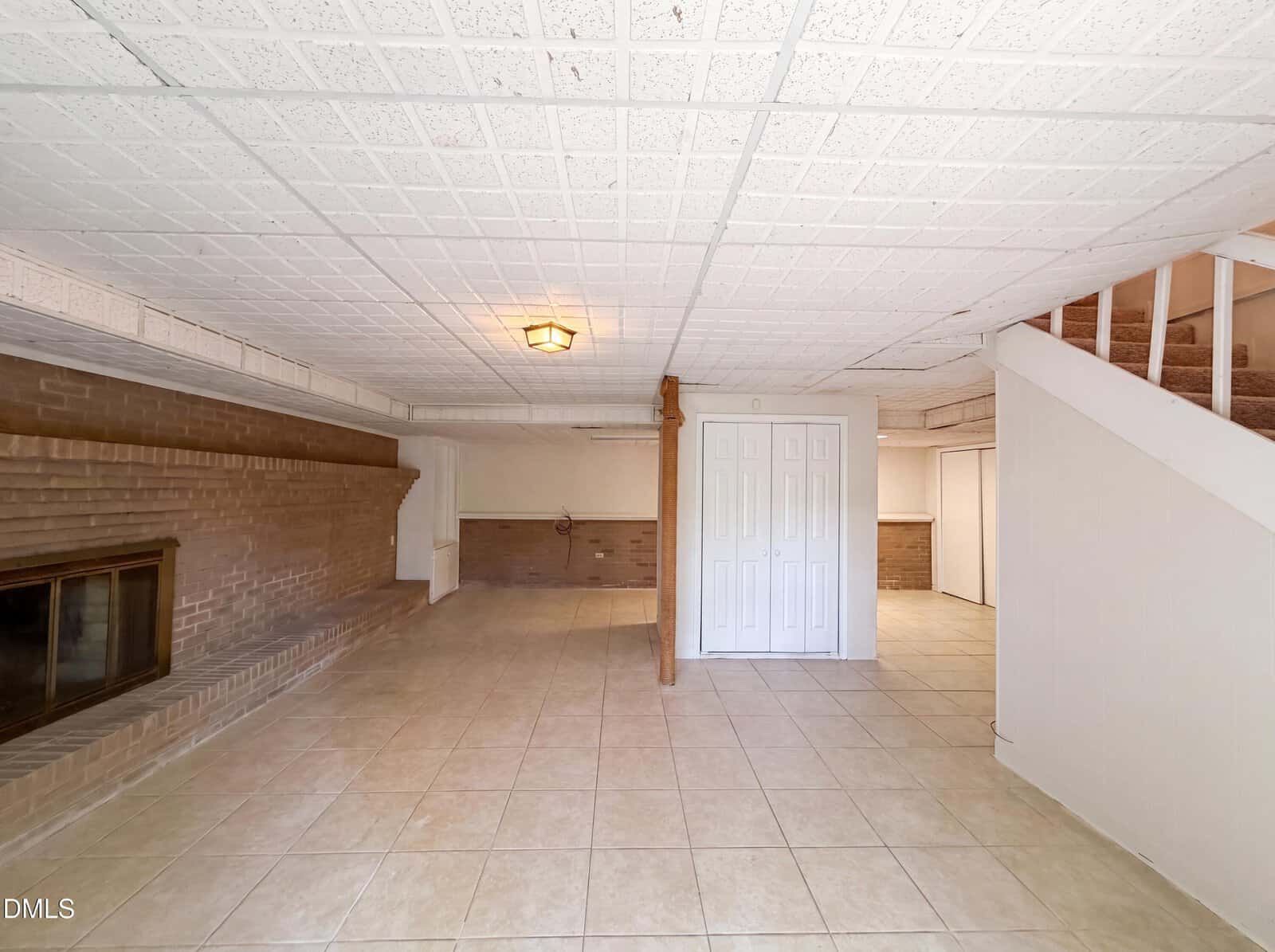 Spacious basement room with tiled floor, brick fireplace on the left, white paneled ceiling, wooden support post, staircase with carpet, and white double closet doors at the back.
