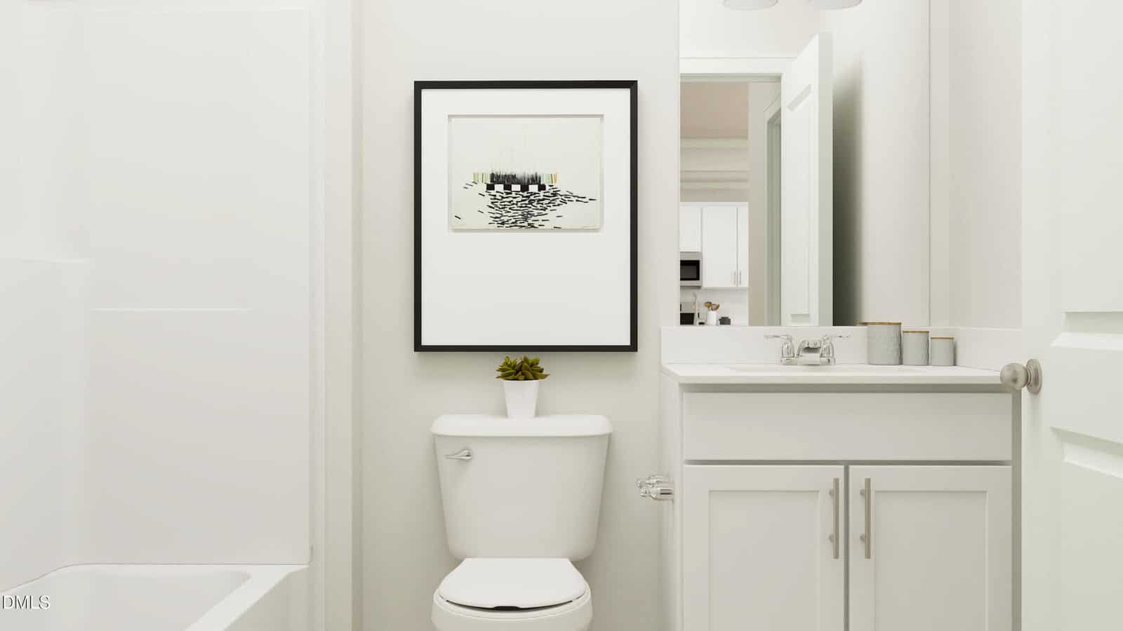 A small white bathroom with a toilet, sink, and bathtub. A framed black-and-white picture hangs above the toilet. A small plant is on the toilet tank, and toiletries are arranged on the sink counter.