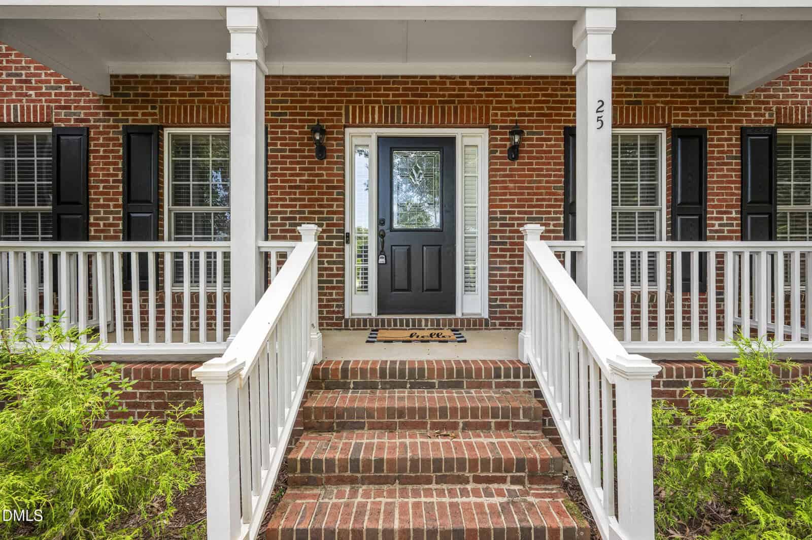 Brick house front porch with white railing, black door, sidelights, house number 25, and welcome mat. Steps lead up to the porch, flanked by green shrubs on each side. Two black light fixtures are by the door.