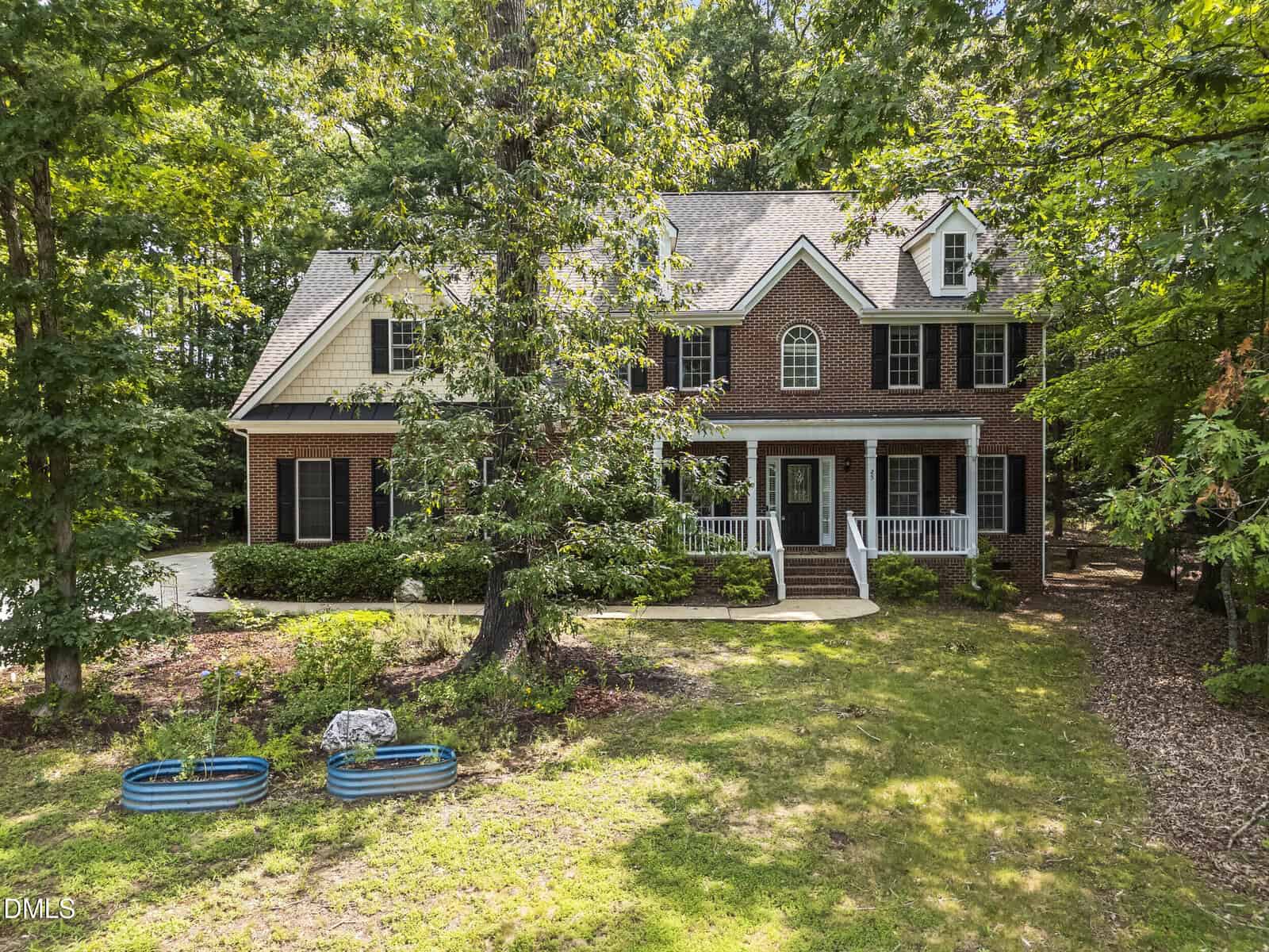 Two-story brick house with white trim, black shutters, and a covered front porch, surrounded by trees and greenery. Two blue kiddie pools are on the lawn in the foreground.