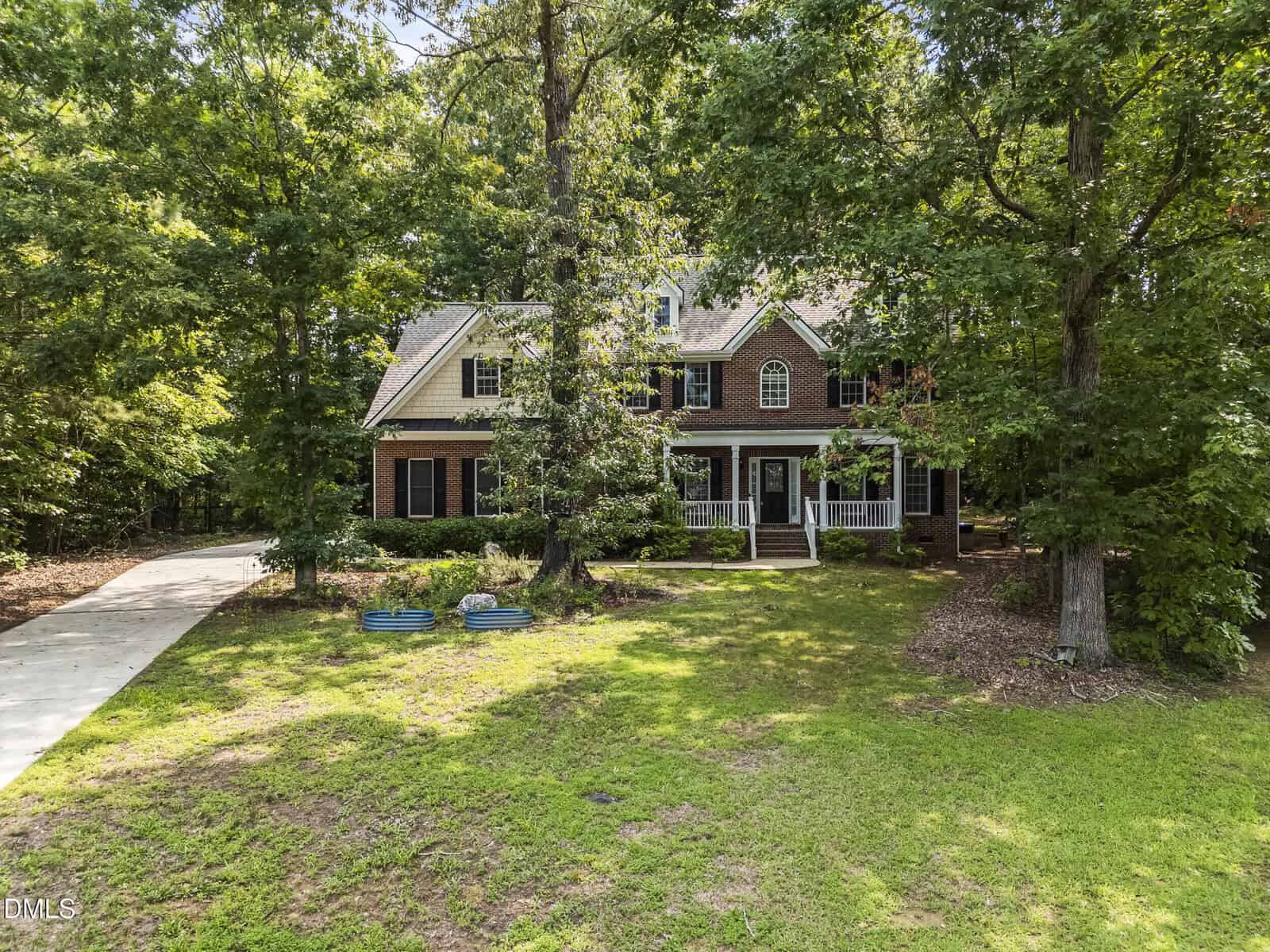 Two-story brick and beige house with white trim, a front porch, and double front doors at 25 Chaucer Court E in Zebulon, surrounded by large trees and a spacious, grassy yard with a driveway on the left.