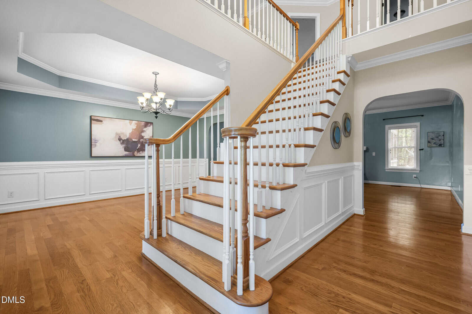 A spacious foyer with a wooden staircase, white railings, hardwood floors, blue-green walls, white wainscoting, and a chandelier above. An adjoining room is visible through an arched doorway.