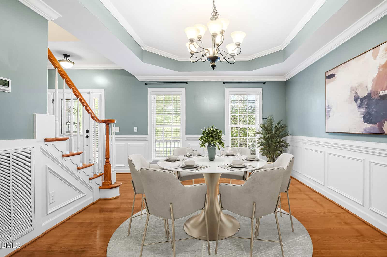 Elegant dining room with light blue walls, wooden floors, a round table set for six, beige chairs, modern art, chandelier, staircase, and large windows with natural light.