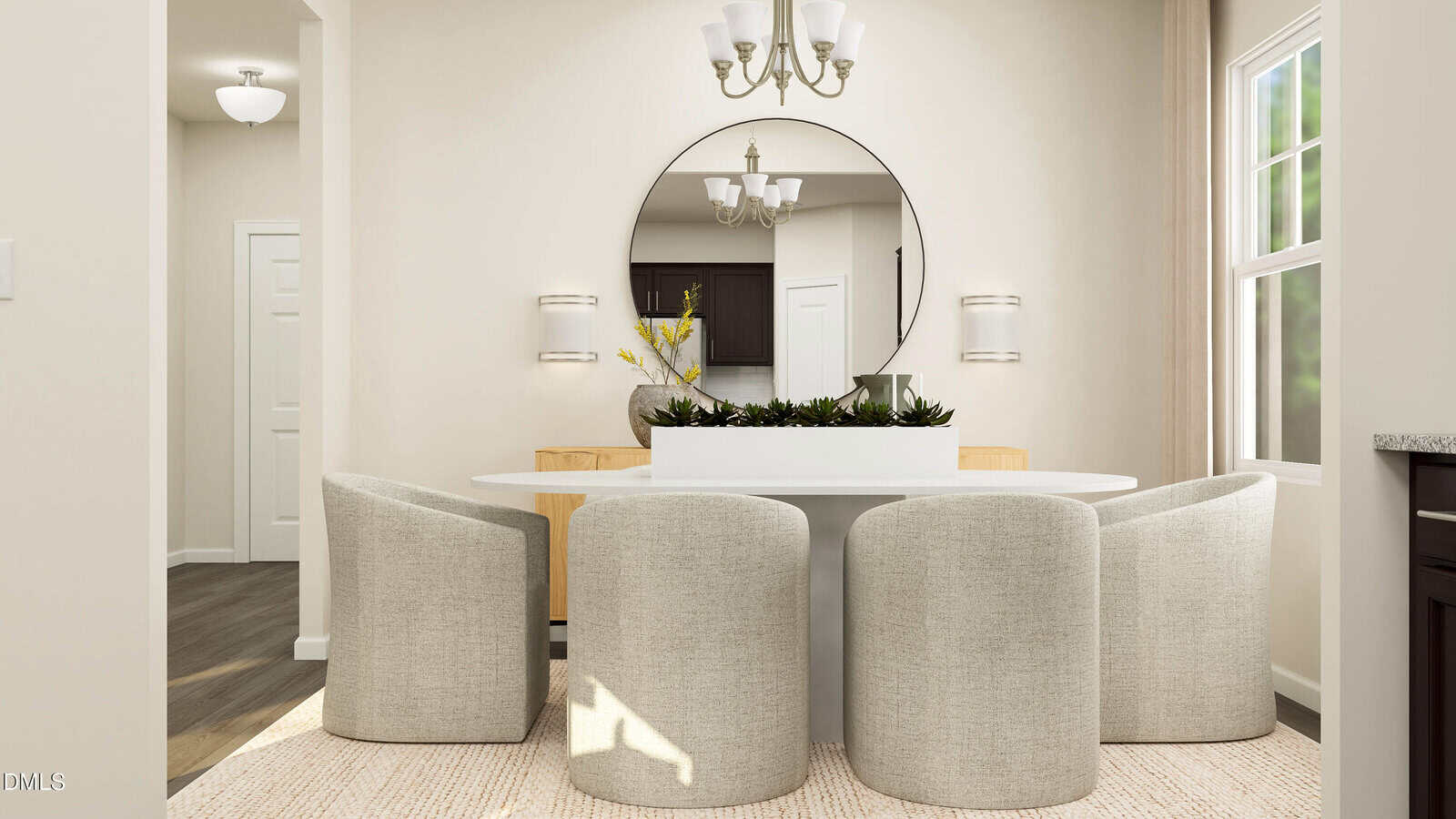 Modern dining room with four light gray upholstered chairs around an oval white table, a large round mirror on the wall, a white planter centerpiece, and two chandeliers overhead. Natural light comes in through a window.