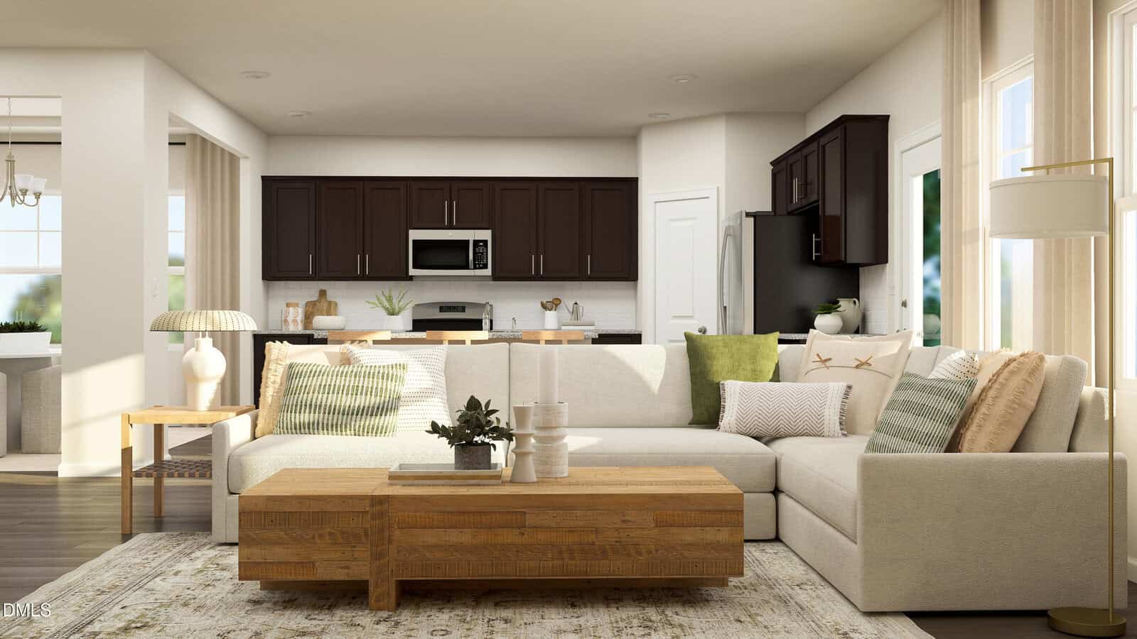 Modern living room with a beige sectional sofa, wood coffee table, and green and white cushions. Open kitchen with dark cabinets and stainless steel appliances is visible in the background. Sunlight streams through large windows.