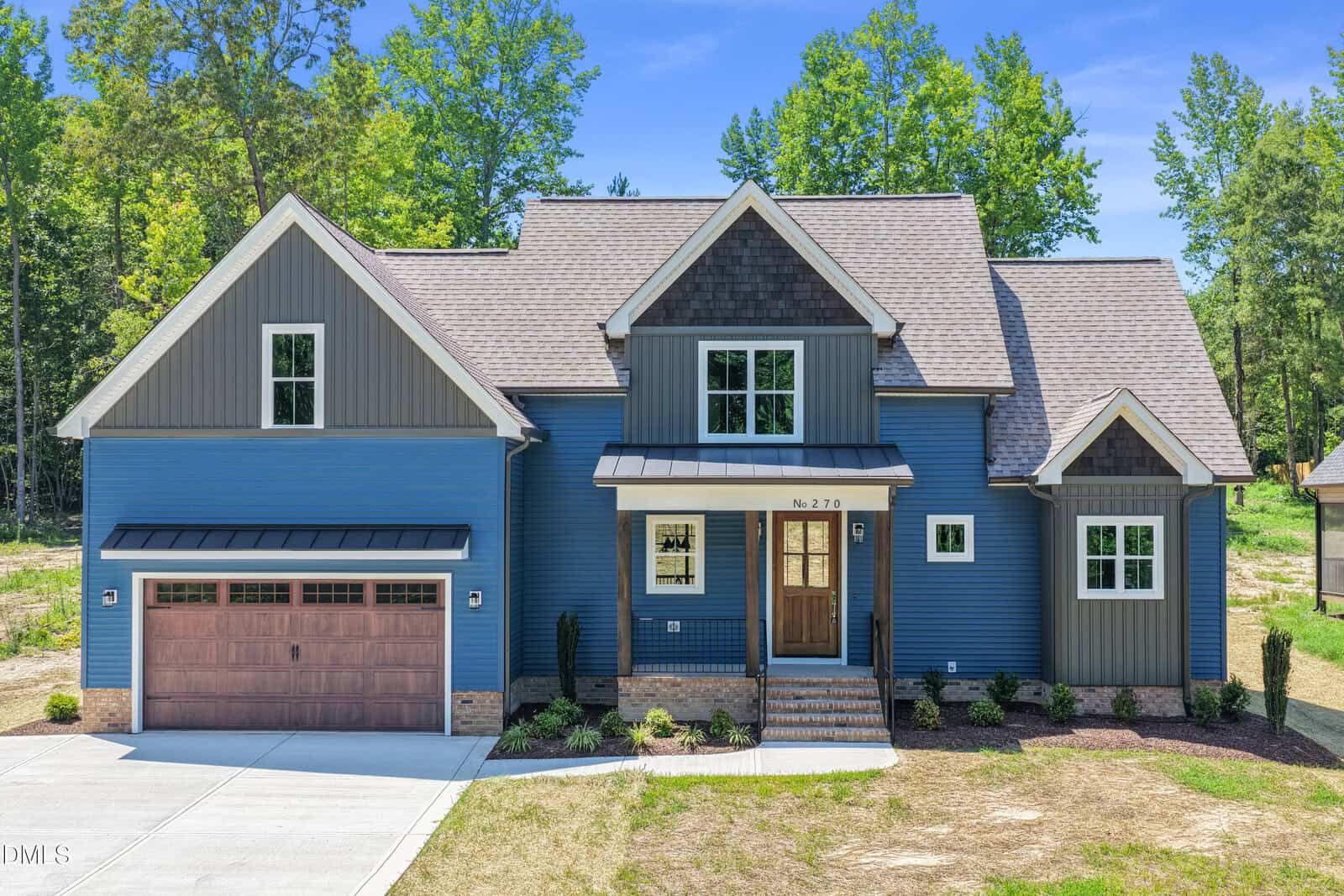 Modern two-story blue house with gabled roof, brick accents, brown double garage door, central front steps, and landscaped front yard on 270 Pilot Ridge Road in Zebulon, surrounded by trees and blue sky in the background.