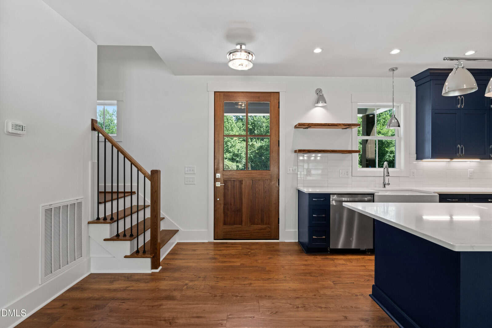 Modern kitchen with dark blue cabinets, white countertops, wood floors, a farmhouse sink, stainless steel appliances, open shelves, and a wooden door with glass panes. Staircase on the left leads to the upper floor.
