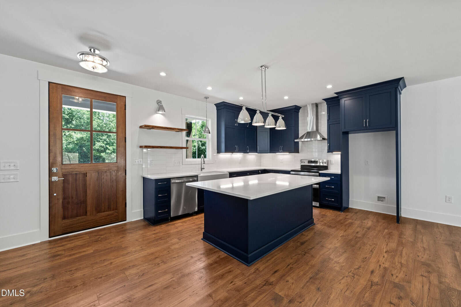 Modern kitchen with navy blue cabinets, white countertops, stainless steel appliances, wooden floors, a central island, open shelves, and a wooden door with a window. Bright, clean, and spacious design.