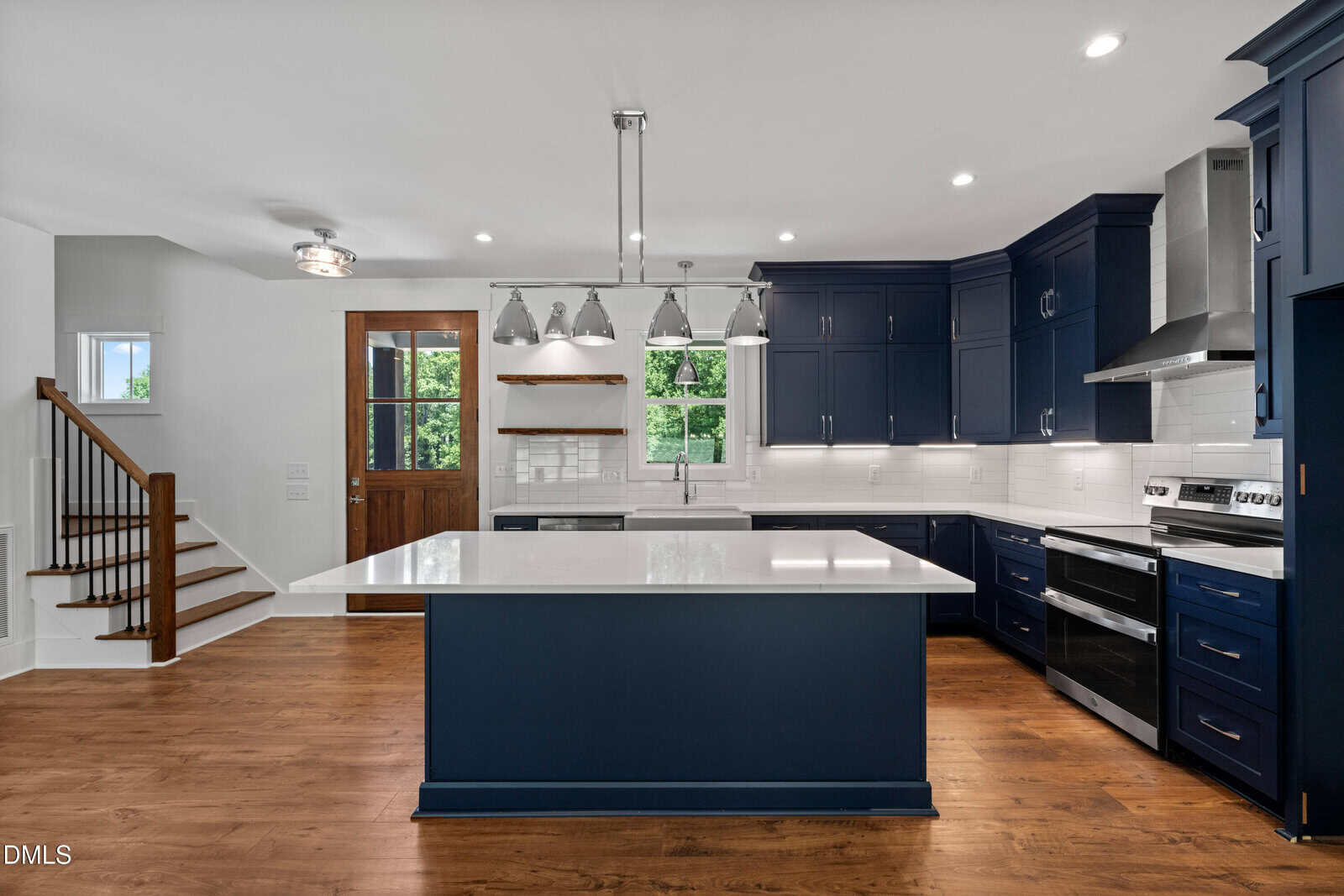 Modern kitchen with navy blue cabinets, white countertops, stainless steel appliances, a large island, wood floors, a wooden door, and a staircase on the left. Bright lighting and a window provide natural light.
