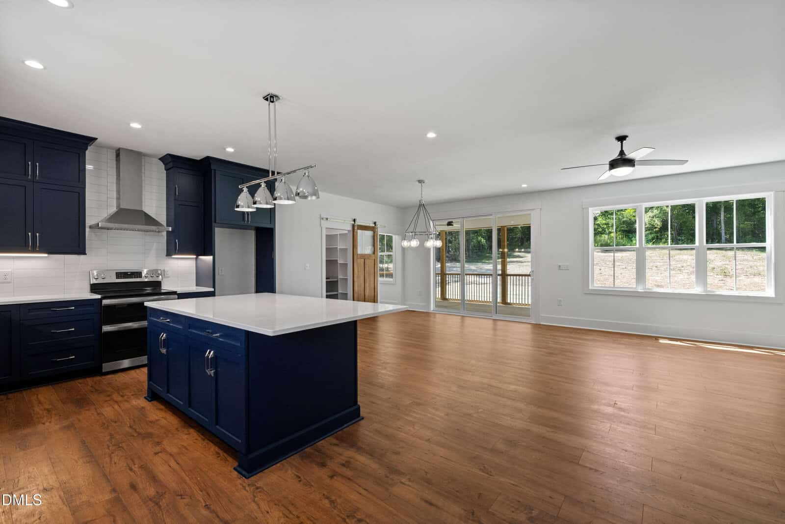 Modern open-concept kitchen and living area with dark blue cabinets, a white island, wood floors, stainless appliances, large windows, ceiling fan, and sliding glass doors leading to a patio.
