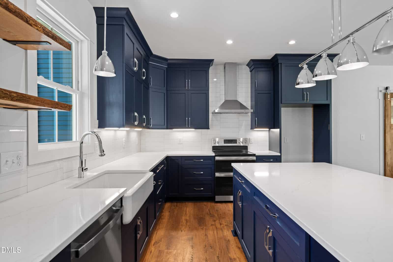 Modern kitchen with navy cabinets, white countertops, stainless steel appliances, farmhouse sink, pendant lights, wooden shelves, and hardwood floors. Barn door visible on the right and white subway tile backsplash behind the stove.