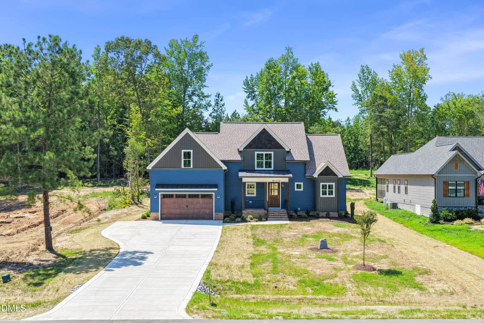 A modern blue house with a two-car garage, front porch, and large driveway, surrounded by trees and neighboring homes under a clear blue sky.
