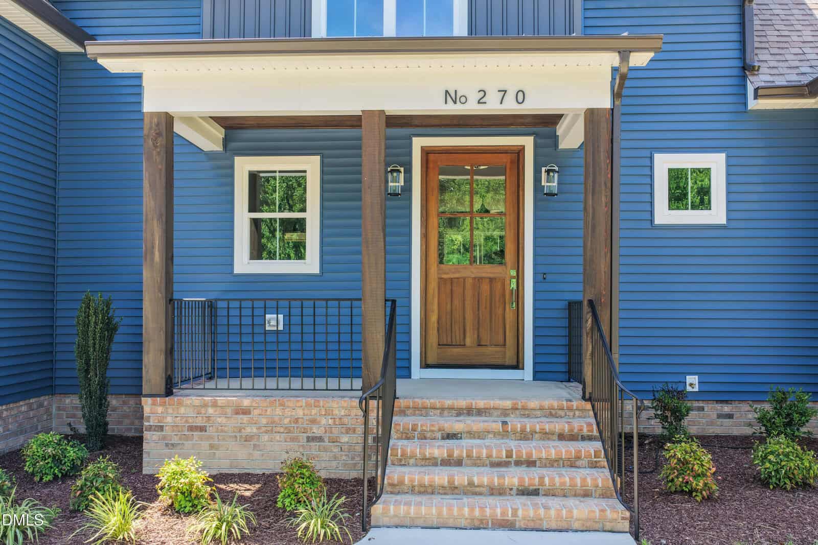Front porch of a blue house with wooden accents, brick steps, black railing, and a wooden front door. The house number above the door reads "No 270." Small shrubs and plants line the entryway.