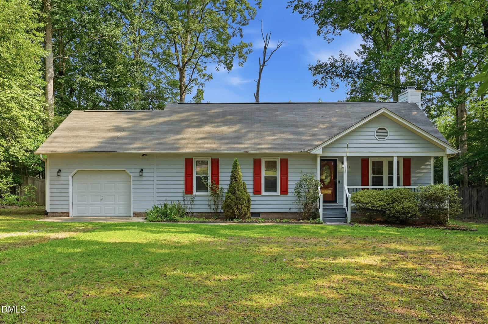 Single-story light blue house with red shutters, a front porch, attached garage, and a green lawn, surrounded by tall trees under a clear blue sky.