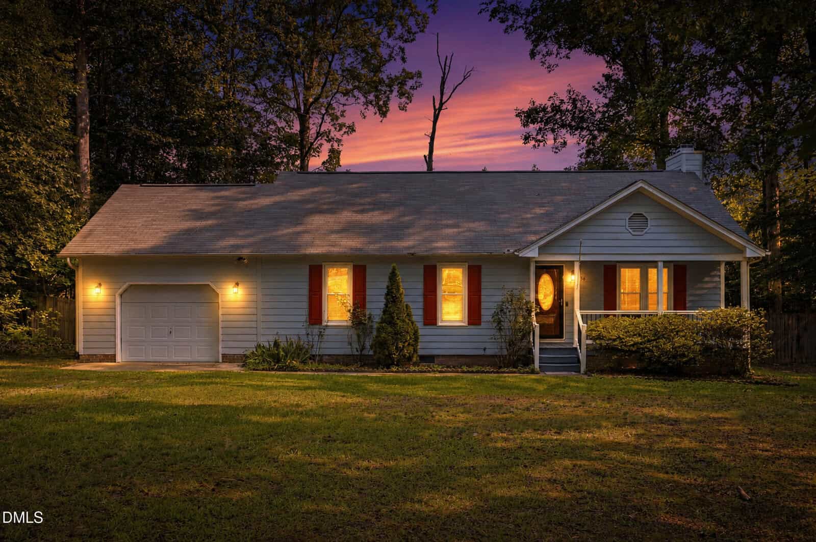 A single-story house with a front porch, garage, and red shutters is warmly lit at dusk, surrounded by trees and a lawn, under a colorful sunset sky.