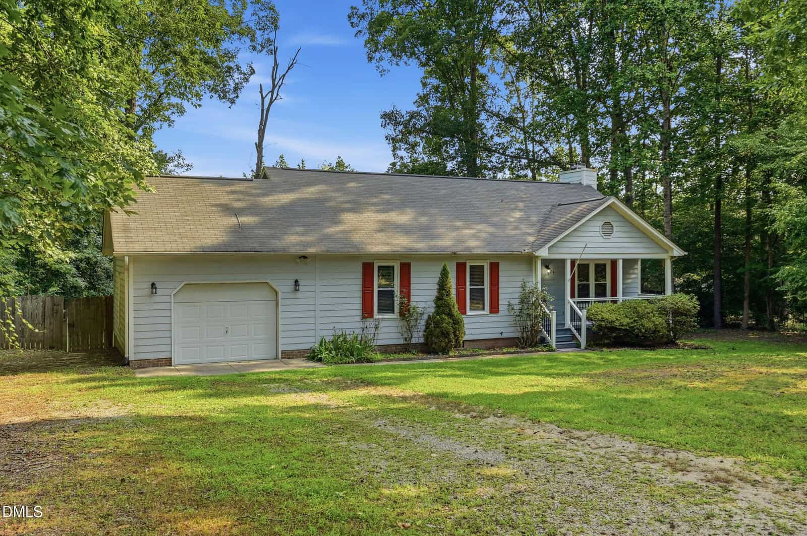 Single-story white house with a one-car garage, red shutters, a small front porch, and surrounded by trees and greenery. There's a gravel driveway and a well-kept lawn in front.