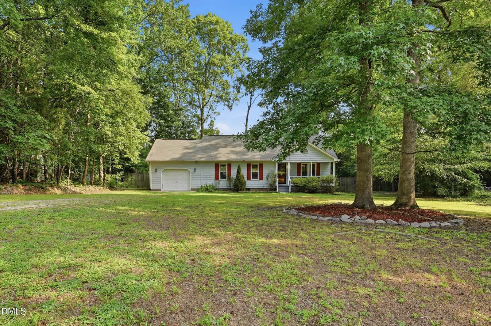 Single-story white house with a porch, red shutters, and attached garage, surrounded by trees and a large grassy yard under a blue sky.