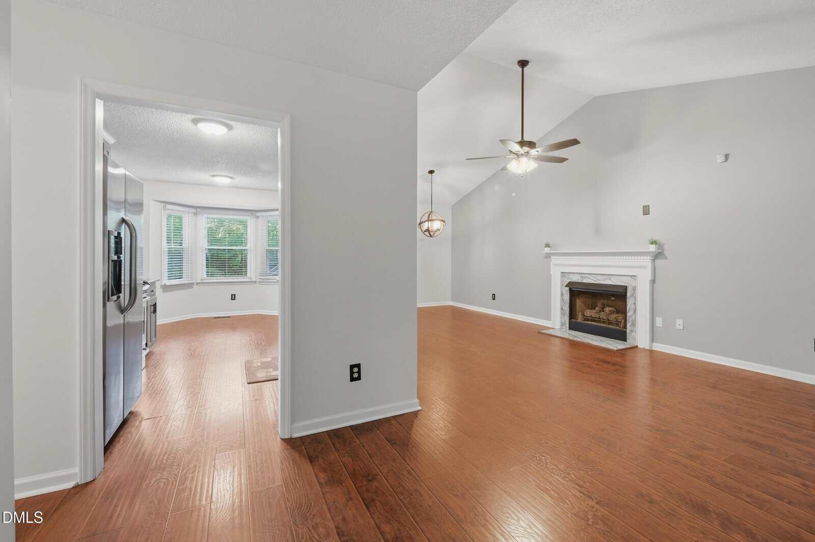 Open kitchen and living room with wood floors, white walls, ceiling fans, a fireplace, and large windows letting in natural light.
