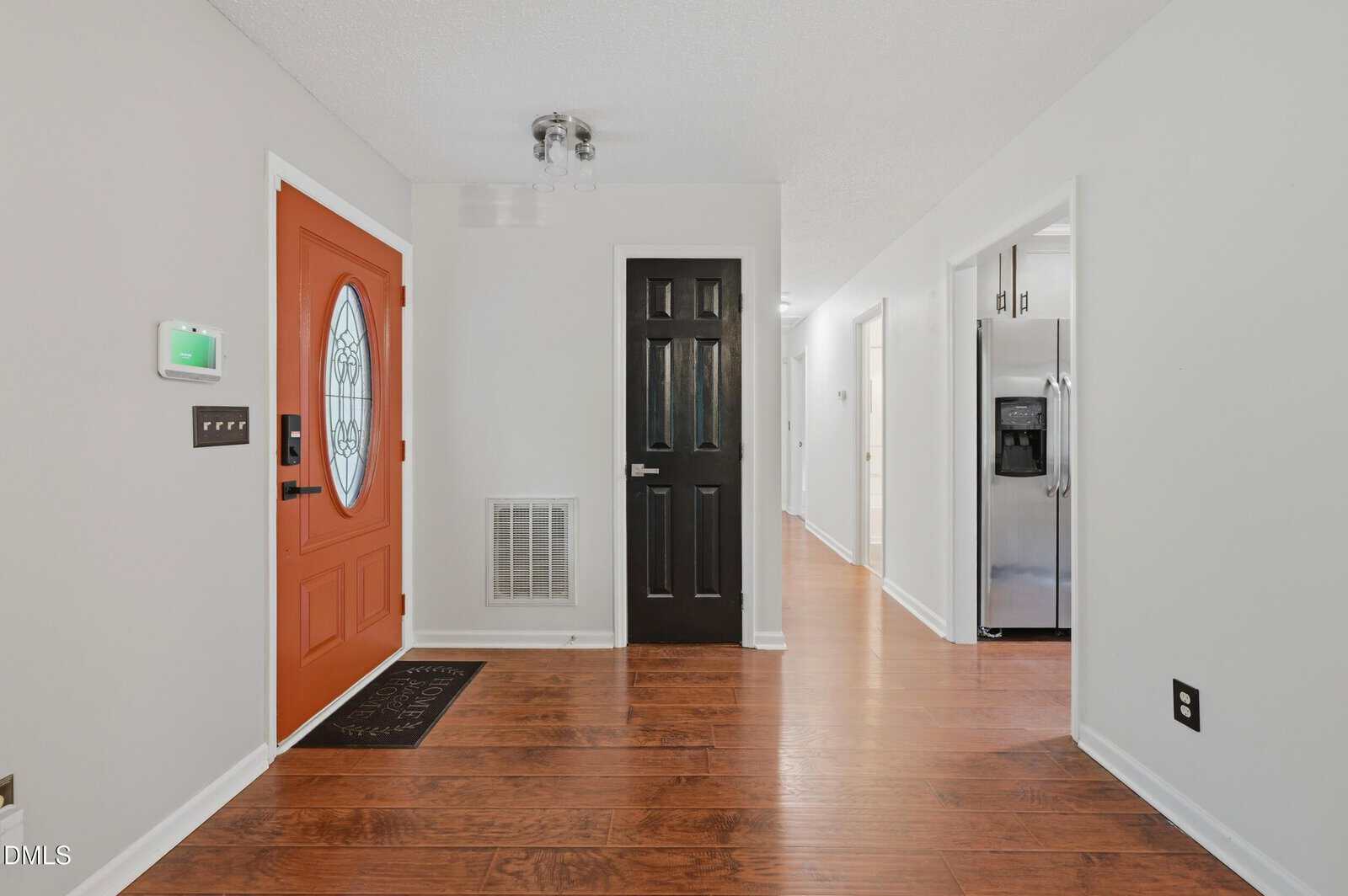 A bright entryway with a red front door, a black interior door, wood flooring, white walls, and a view into a hallway and kitchen with stainless steel appliances.