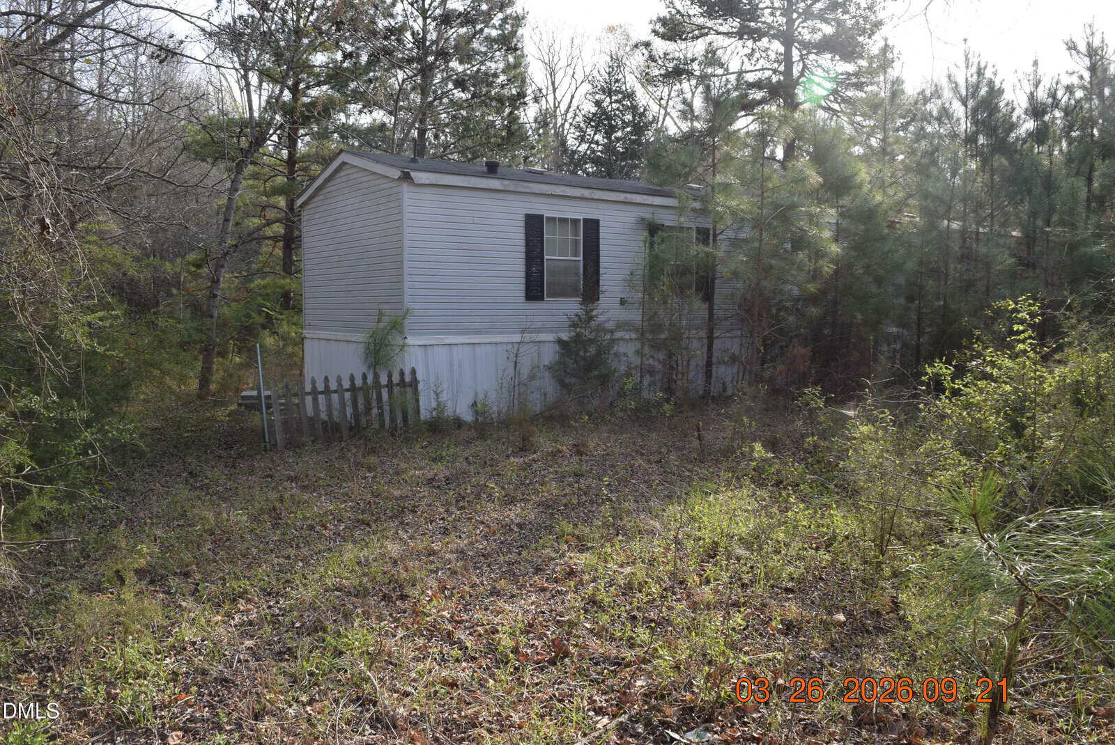 A small white mobile home sits in an overgrown yard surrounded by trees and bushes, with a broken wooden fence visible on the left at 309 Bluegrass Drive in Warrenton.