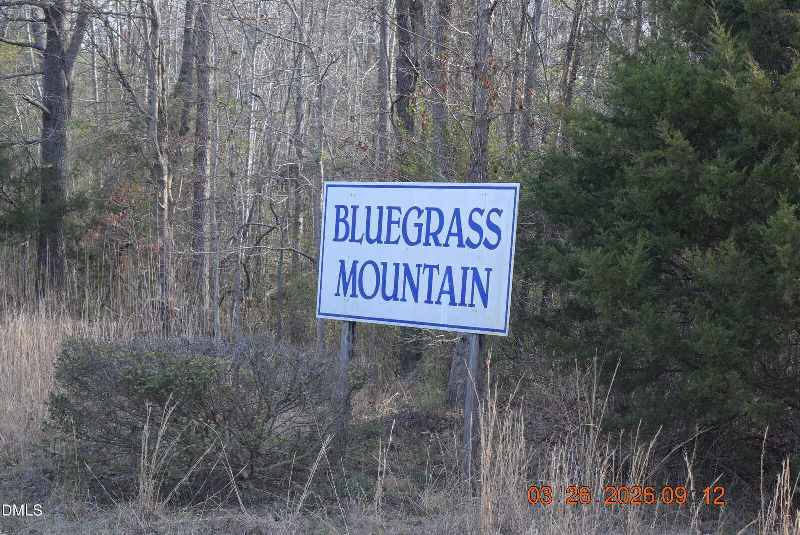 A white sign with blue letters reads “BLUEGRASS MOUNTAIN.” It stands amid grass and bushes with trees in the background.