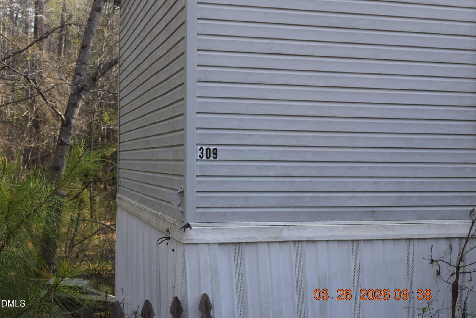 The corner of a white mobile home with the number 309 on the side. Trees and greenery are visible in the background. The date “03-26-2026” appears in orange at the bottom right.