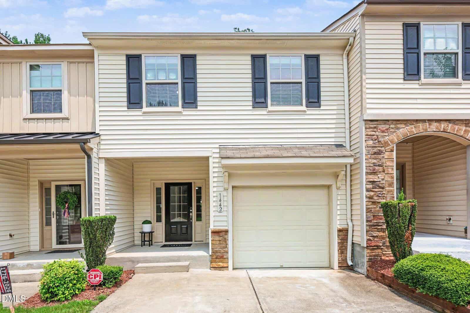 Located at 1442 Montonia Street in Wake Forest, this beige two-story townhouse features black shutters, a single-car garage, a small covered porch, neatly trimmed shrubs, and a concrete driveway leading to the front entrance.