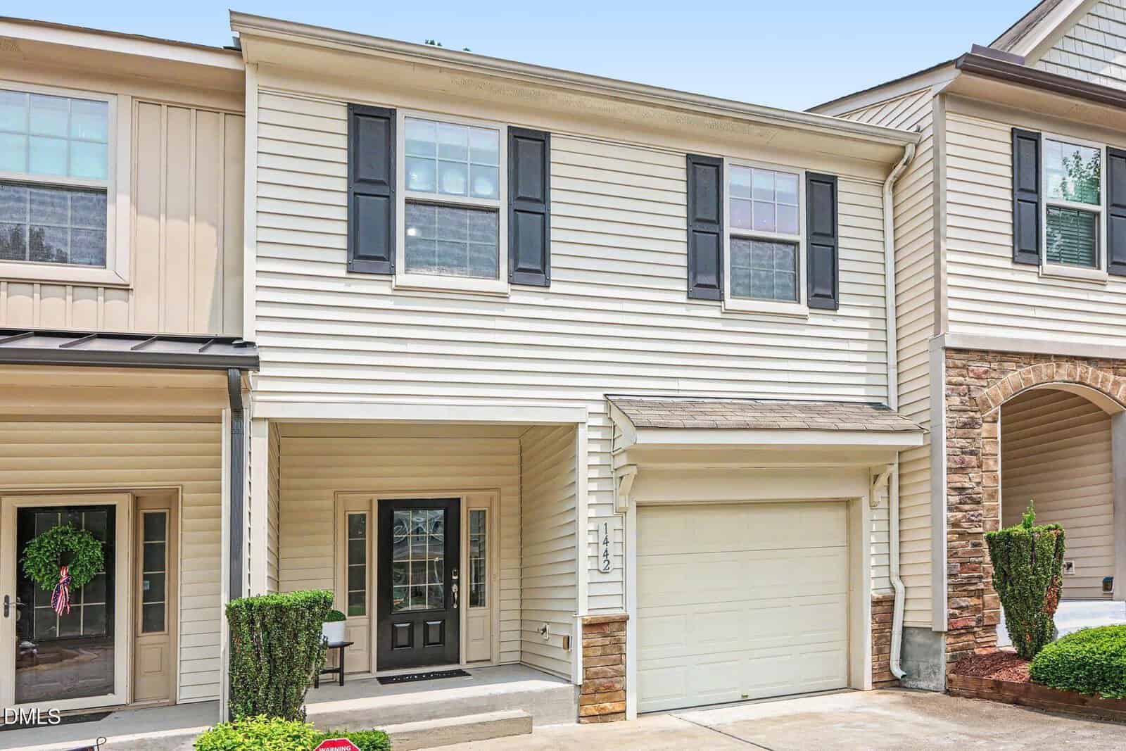 Two-story townhouse with beige siding, black shutters, a black front door, single-car garage, and neatly trimmed bushes by the entrance on a sunny day.