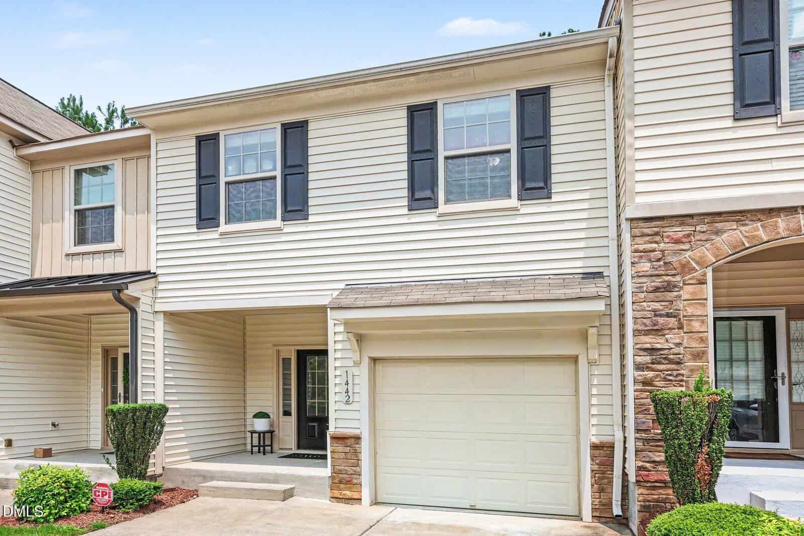 Two-story beige townhouse with black shutters, a single-car garage, front porch, and neatly trimmed shrubs along the driveway and entrance.