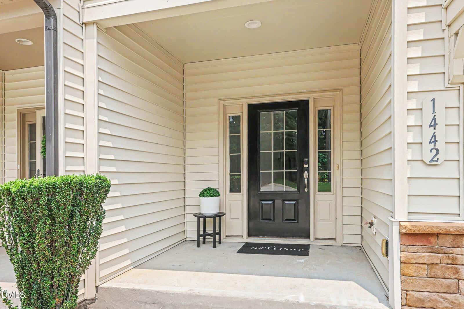 Covered porch with cream-colored siding, black front door with glass panels, a welcome mat, potted plant on a small white table, house number 1442 on the wall, and neatly trimmed bush visible on the left.