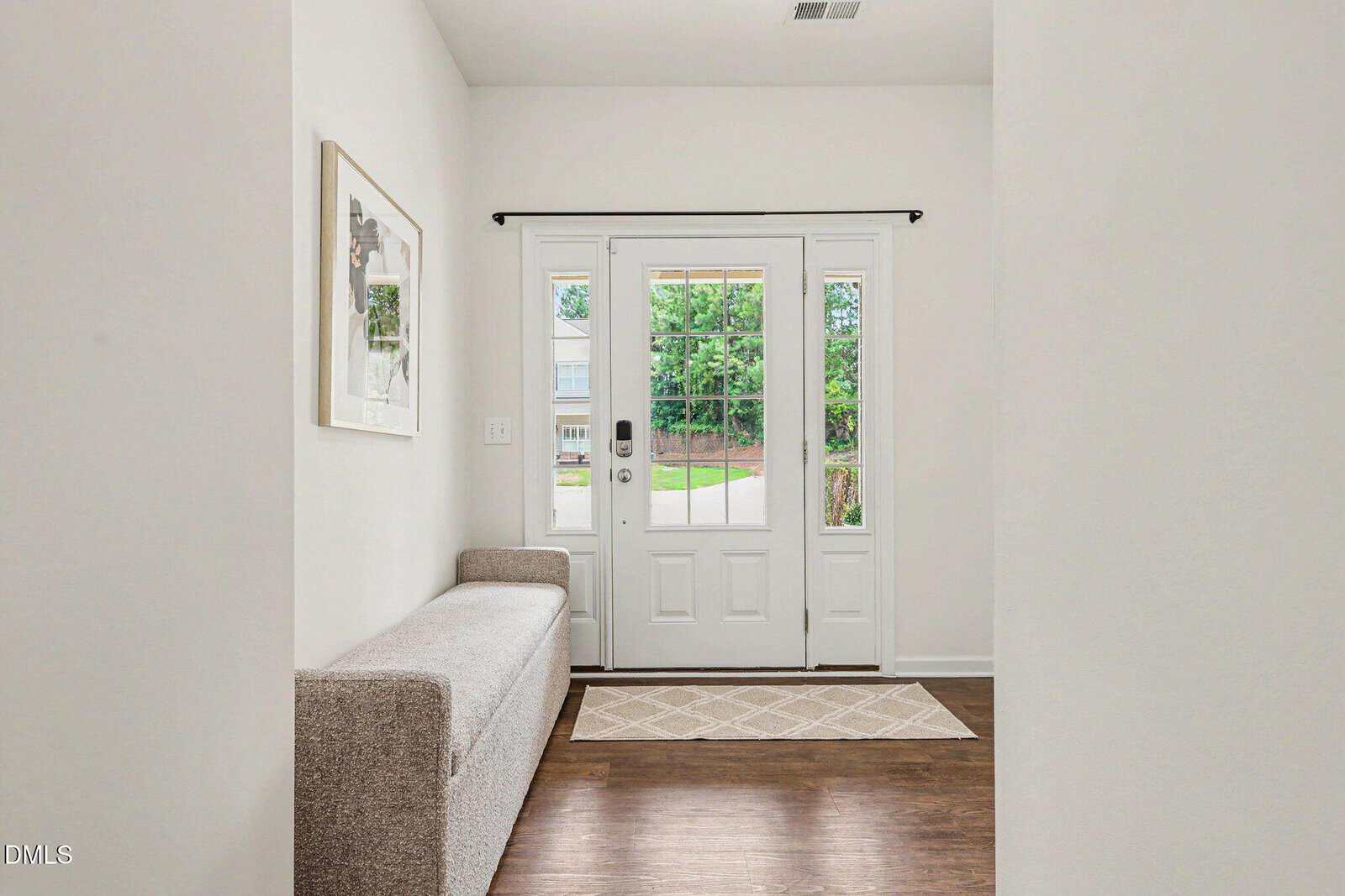 A bright entryway with a white front door, glass side panels, a beige rug, a cushioned bench on the left, and a framed art piece on the wall. Hardwood floors and a view of the outdoors are visible.