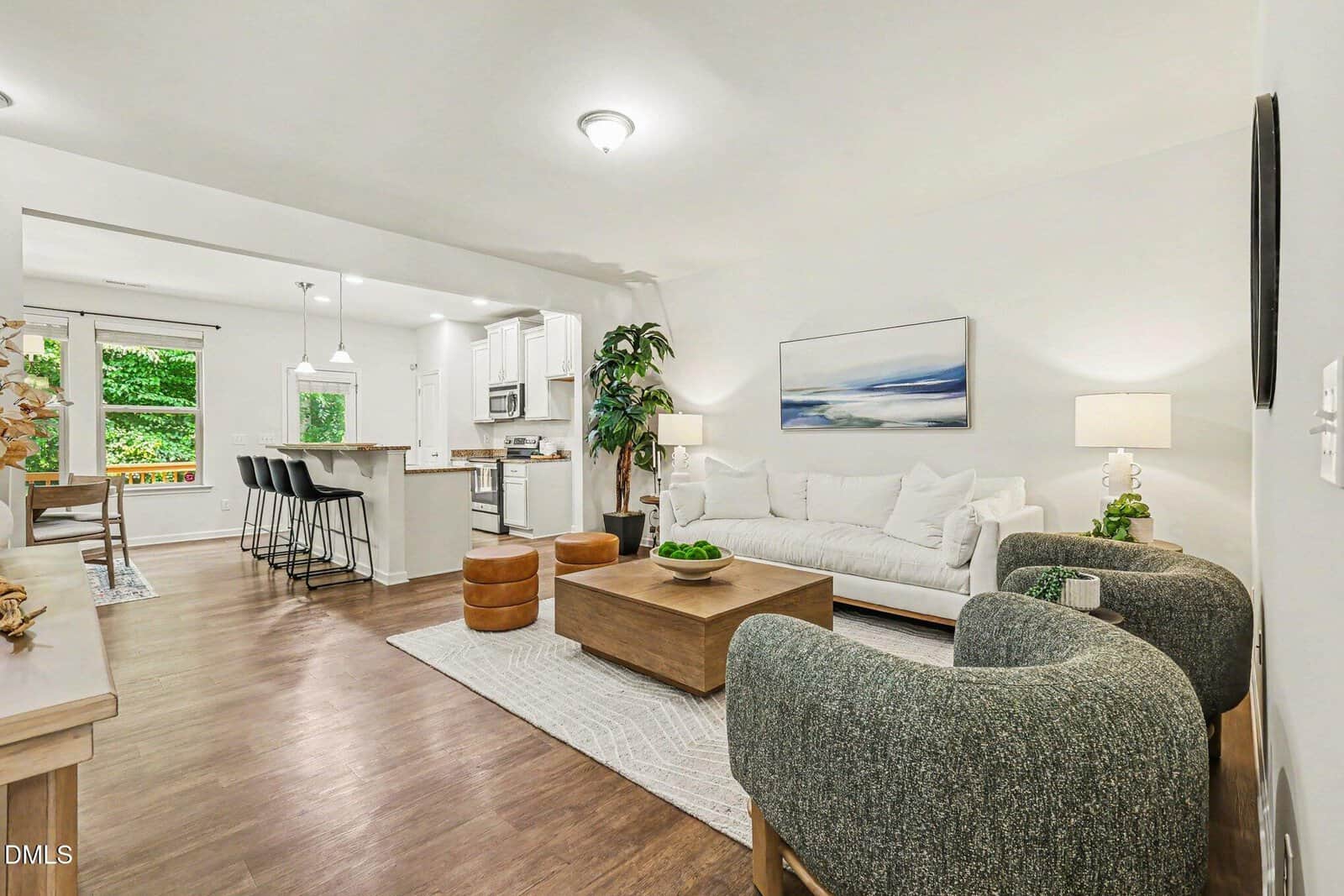 Bright, modern living room with a white sofa, two gray chairs, wood coffee table, kitchen with island, barstools, large window, and greenery. Artwork and lamps add cozy, stylish touches to the open space.
