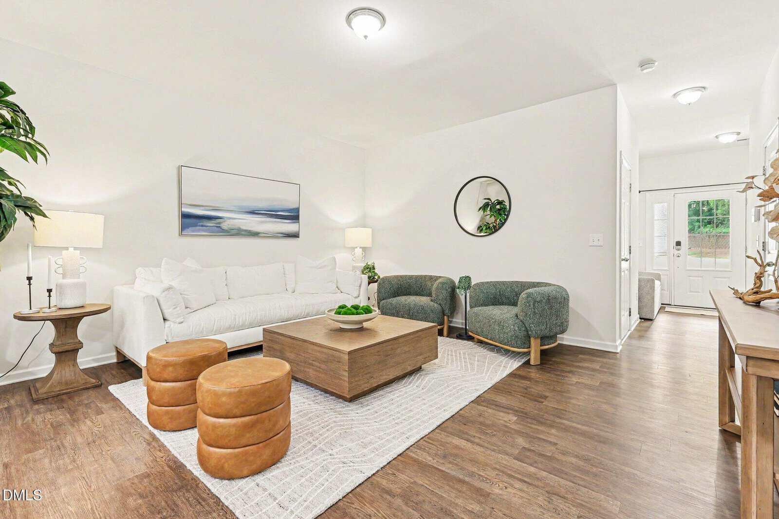 Bright living room with a white sofa, two green chairs, round mirror, wooden coffee table, two brown ottomans, and a large rug on wood floors. Light walls and modern decor create a cozy, welcoming space.